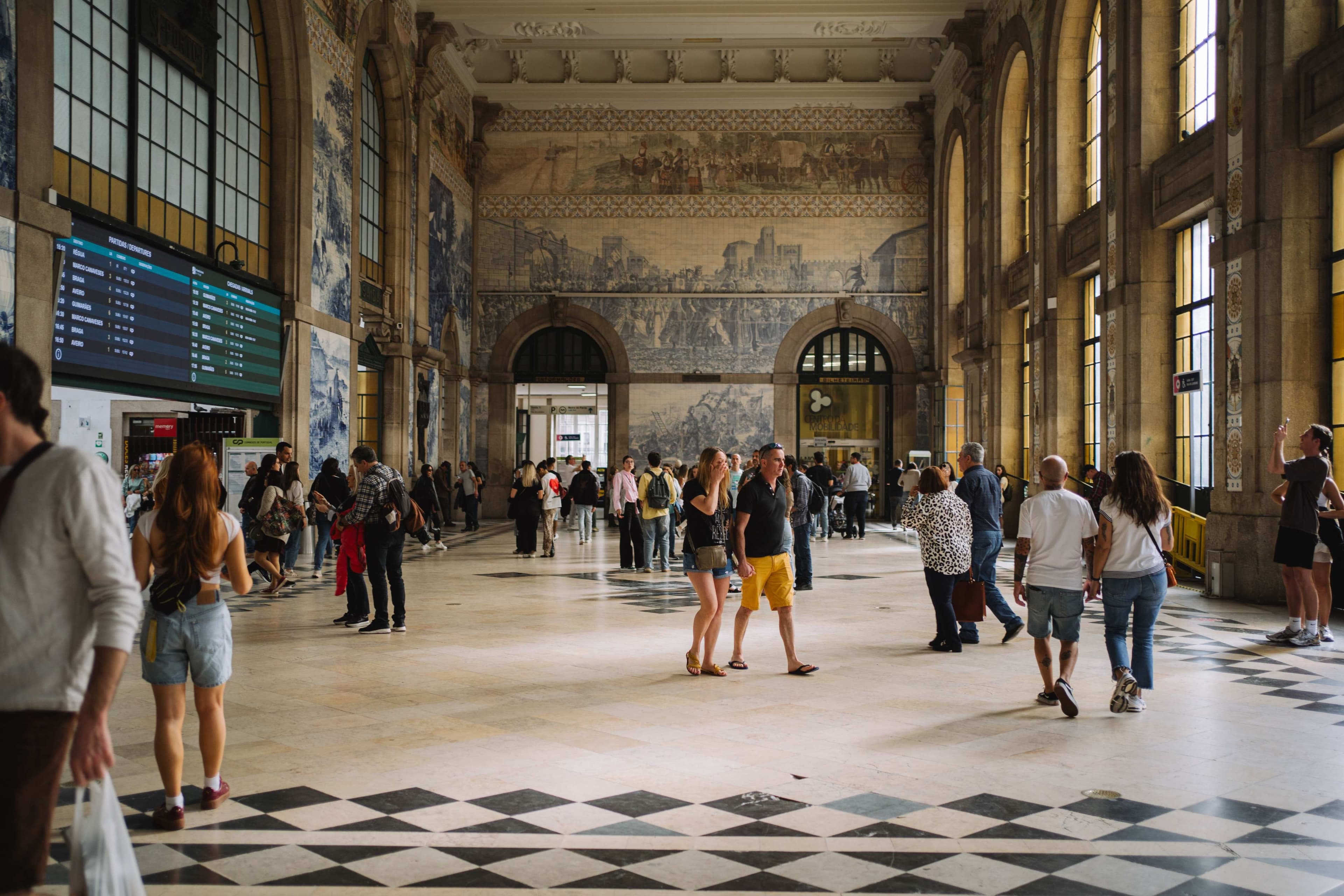 Pessoas no átrio de uma estação de comboios, com grandes painéis de azulejos nas paredes e janelas altas.